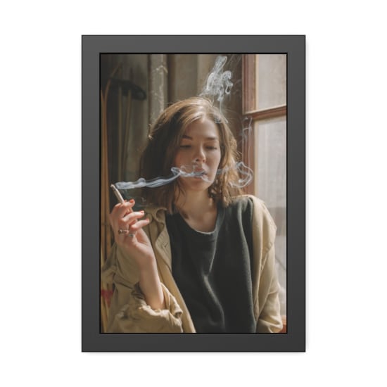 Thoughtful woman in casual attire smoking indoors with sunlight streaming through the window.