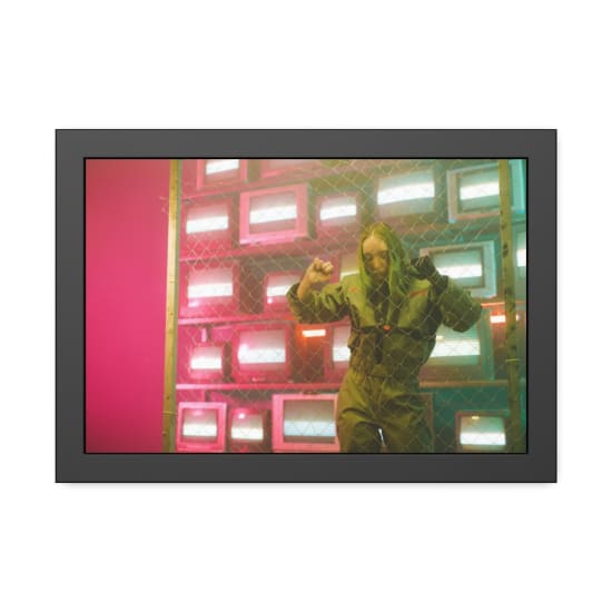 A woman in a jumpsuit stands behind a wire fence in a room filled with glowing TV screens.