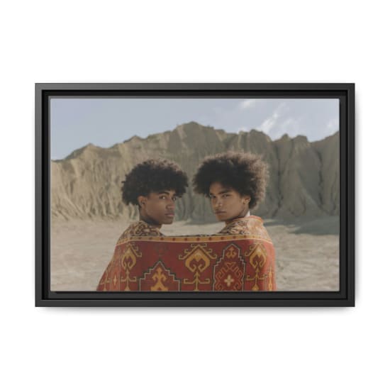 Two young men with afro hairstyles look back, wrapped in a patterned blanket in a desert landscape.