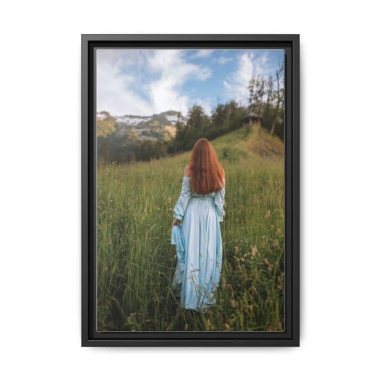 Redhead woman in a flowing light blue dress walks through a lush green field with mountains in the background.