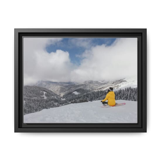 Snowboarder in yellow jacket sitting on snow, admiring a snowy mountain landscape under a cloudy sky.