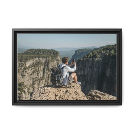 A man sits on a cliff edge taking a photo of a scenic canyon with his phone, capturing a breathtaking landscape.