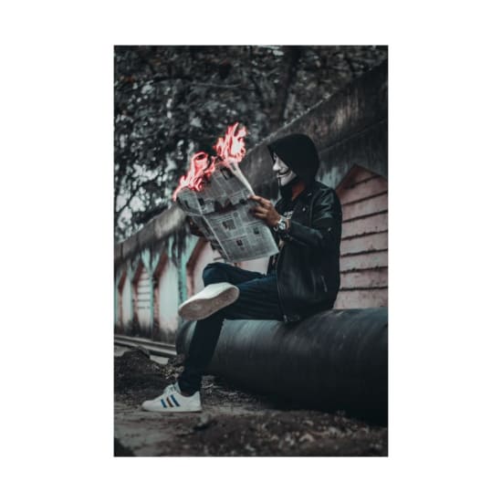 A person wearing a mask reads a flaming newspaper on an urban street, creating a surreal and mysterious scene.