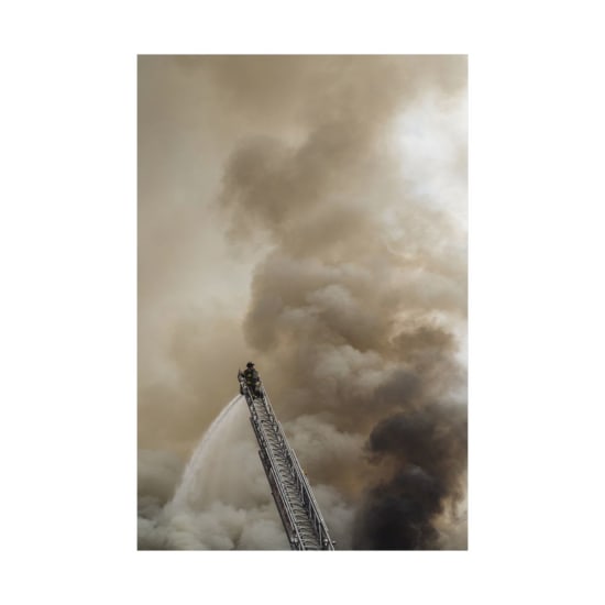 Brave firefighter extinguishing fire from a ladder amidst heavy smoke backdrop.