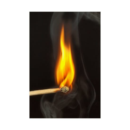 A detailed view of a matchstick burning with bright flame against a dark background, emitting smoke.
