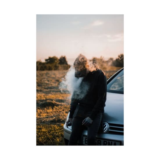 A man stands relaxed, vaping beside a car in a golden-hour field setting.