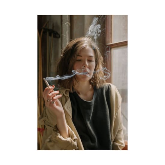 Thoughtful woman in casual attire smoking indoors with sunlight streaming through the window.