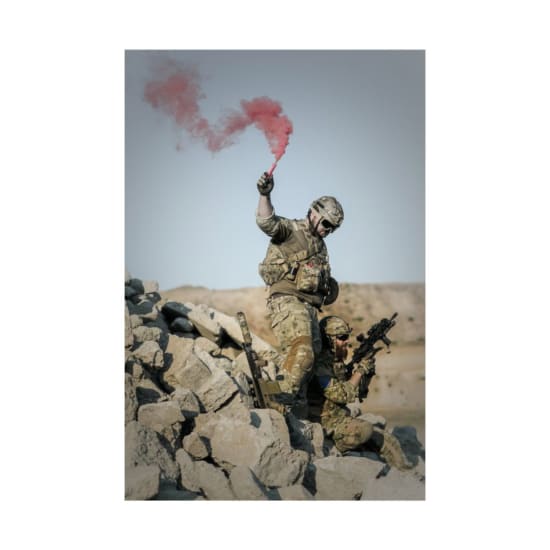 Soldiers in camouflage conduct a military operation using a red smoke signal.