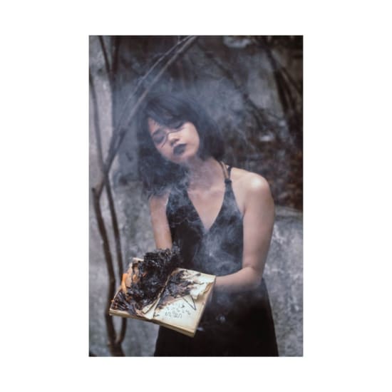 An artistic photo of a woman in a black dress holding a burning book amidst smoke.