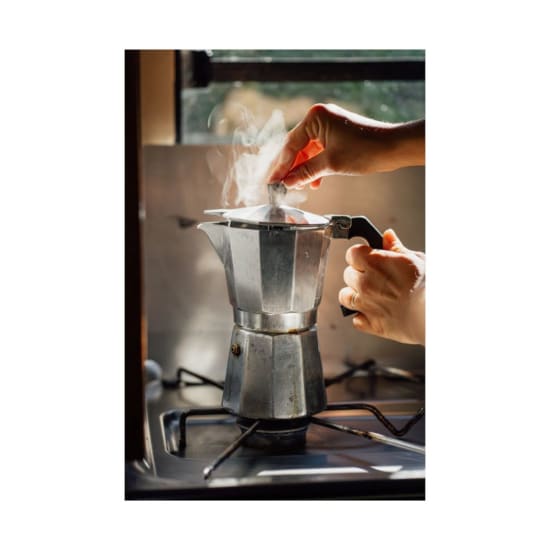 Close-up of hands operating a stovetop espresso maker with visible steam in a cozy kitchen setting.