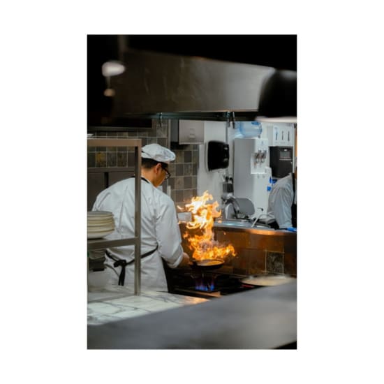 A chef skillfully handles a flaming pan in a commercial kitchen, showcasing culinary expertise.
