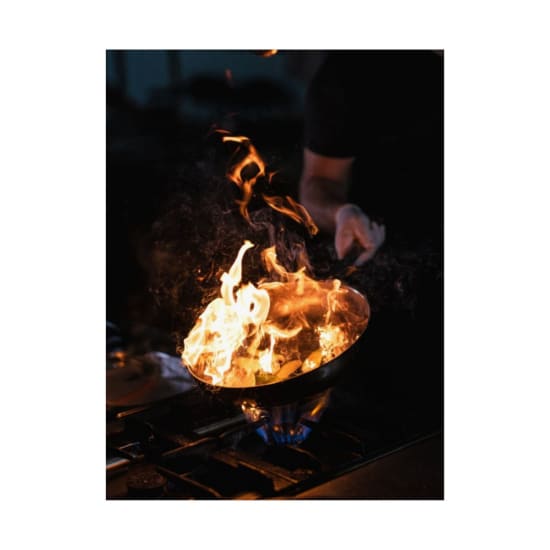 A chef skillfully maneuvers a pan engulfed in vibrant flames on a stove.