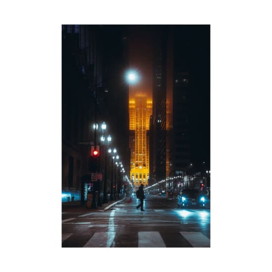 A lone figure crosses a city street at night, illuminated by a glowing skyscraper.