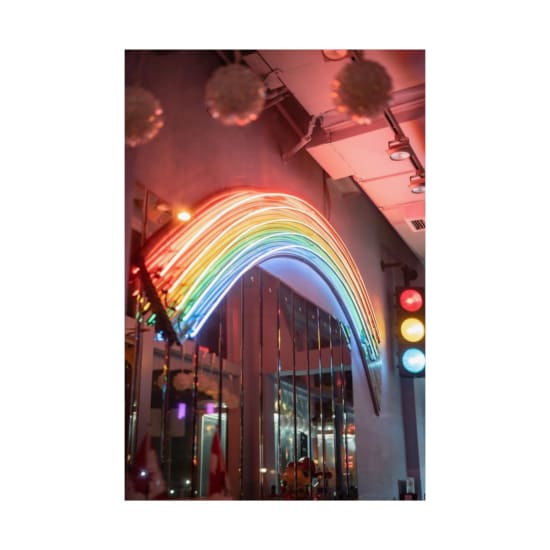 A colorful neon rainbow arch illuminates a bustling urban environment at night.