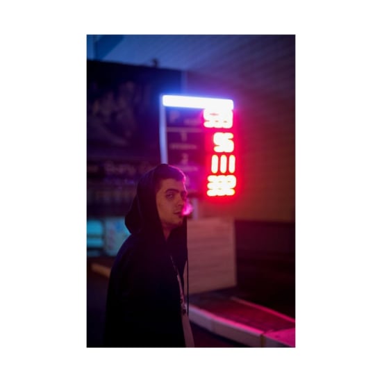 A man in a hoodie stands illuminated by neon lights, exhaling smoke at night.