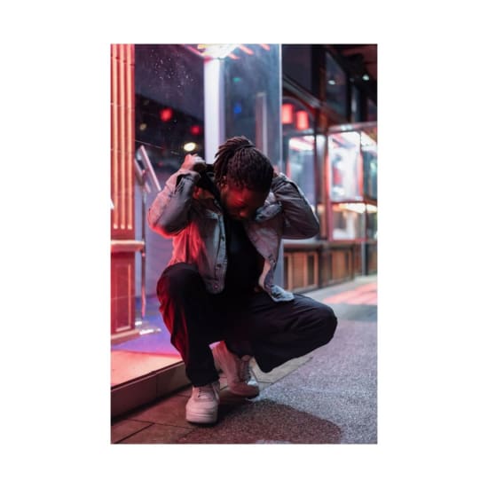 Full body of young African American male with dreadlocks sitting on haunches on street and putting on hood against luminous lights at night