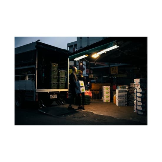 Workers unloading boxes from a delivery truck at an urban market during dusk.