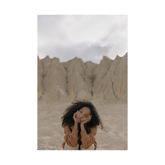 A fashionable young woman with curly hair poses thoughtfully in a striking desert landscape.