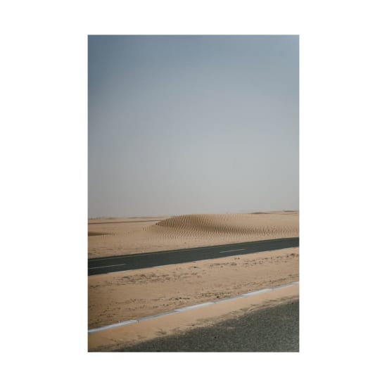 A remote asphalt road cuts through a sandy desert with dunes under a clear sky.