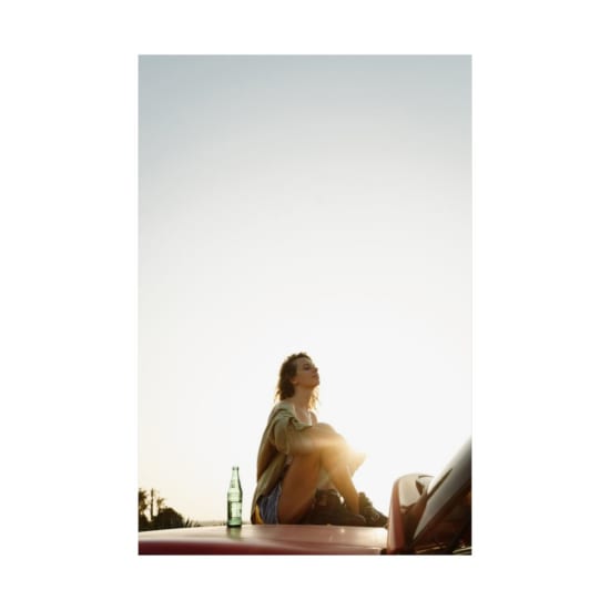 Young woman sitting on a car under bright sunlight, enjoying a peaceful moment outdoors.