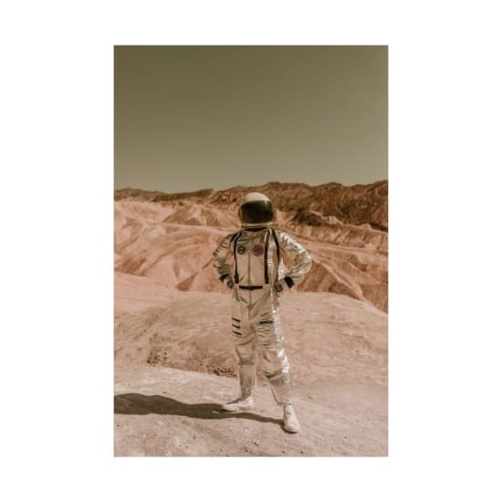 A person in a space suit stands confidently in a barren, desert-like landscape, evoking exploration of distant planets.