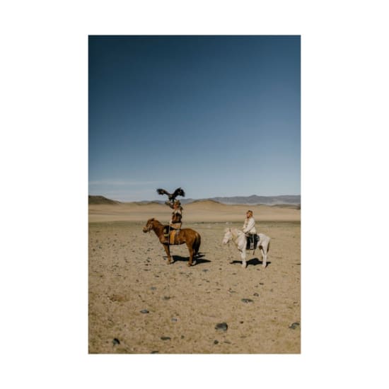 Side view of ethnic people in traditional clothes riding horses in dry sandy desert covered with pebbles