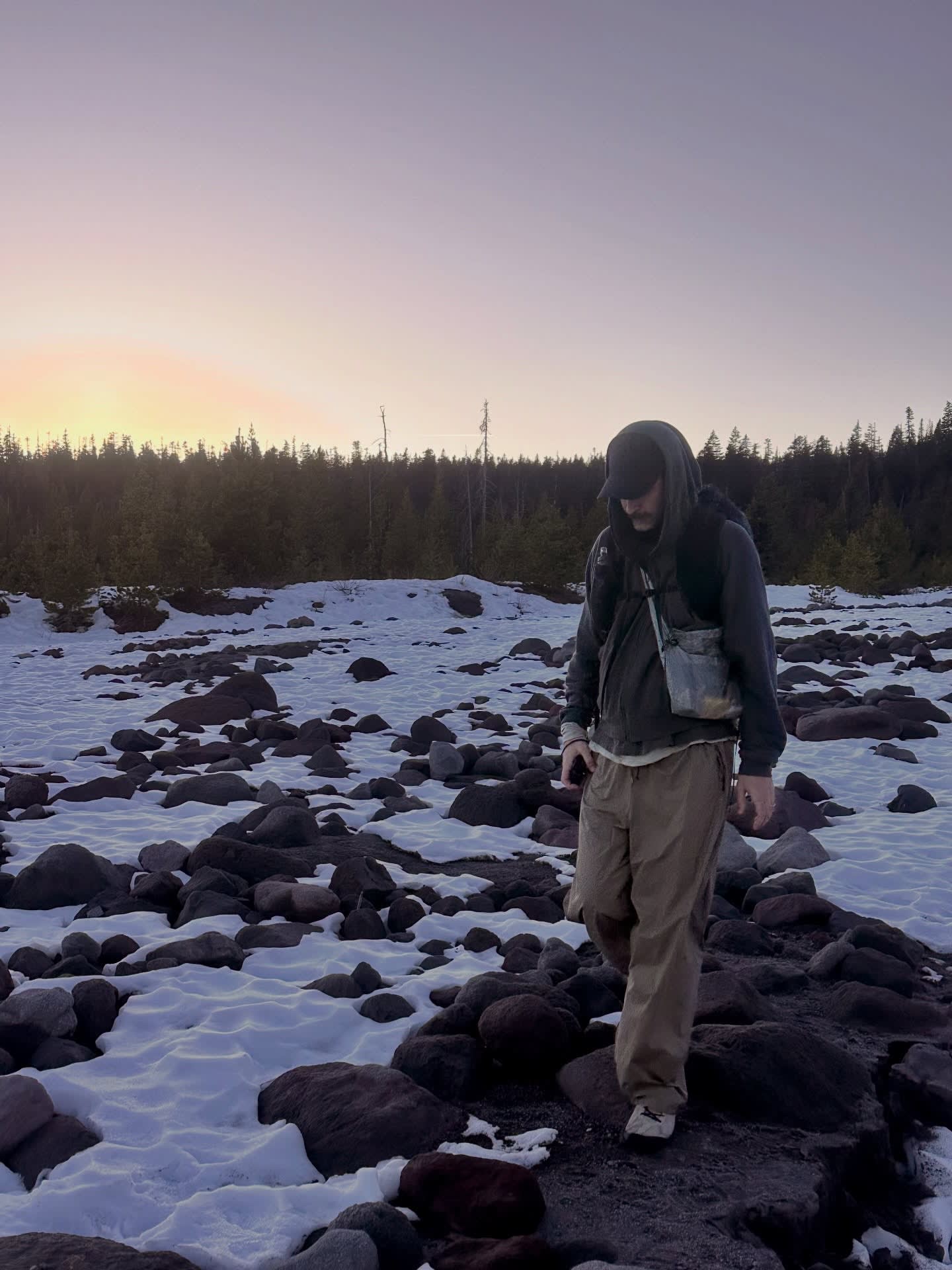 A person in a hooded jacket and tan pants stands on a snowy, rocky terrain against a backdrop of a distant forest and a setting sun.