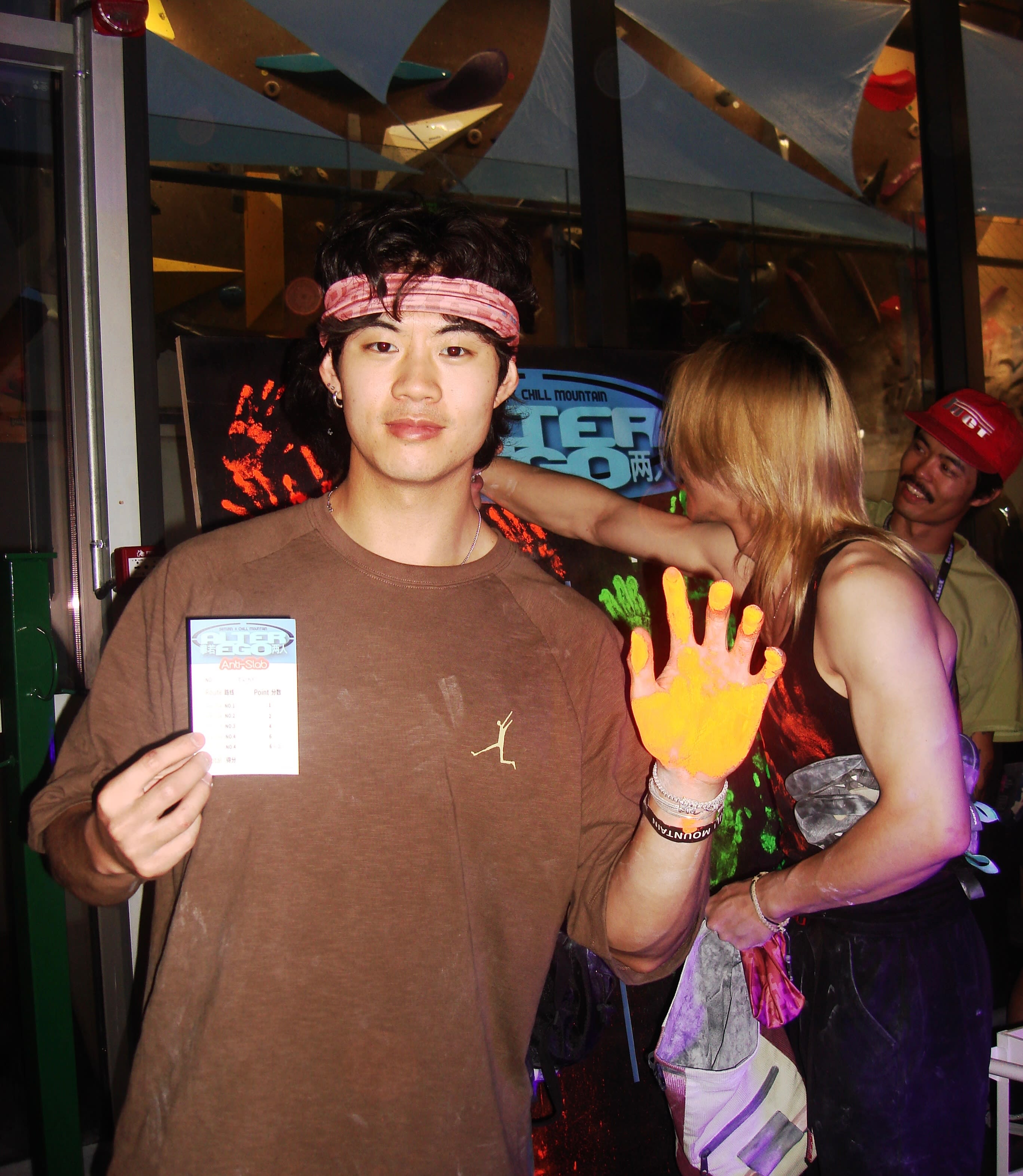 A young man in a brown t-shirt and headband holds a small card in the foreground, while a person with yellow paint on their hand and another man in a red cap stand behind him.