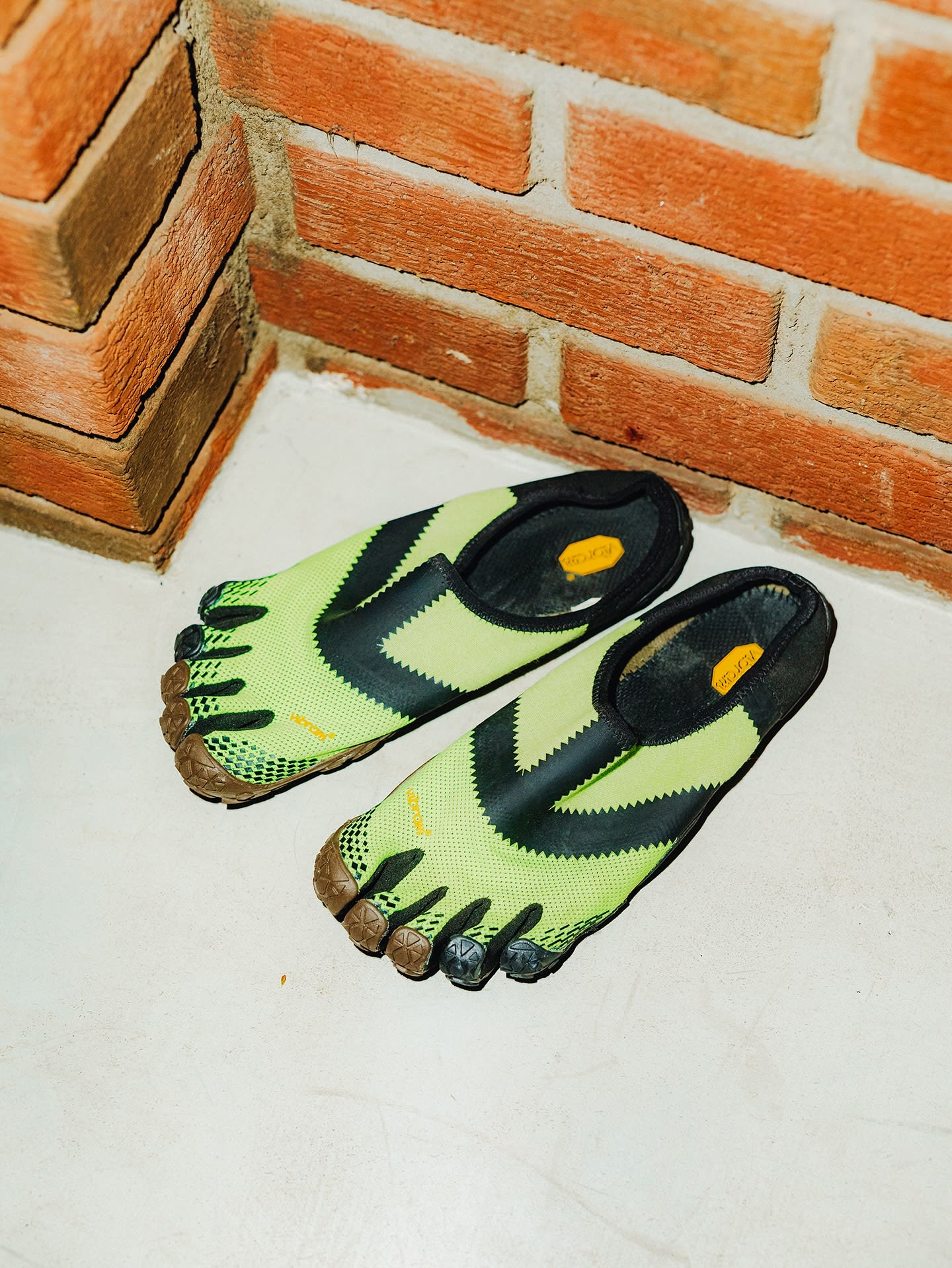 A pair of lime green and black five-finger shoes sit on a white floor against a red brick wall.
