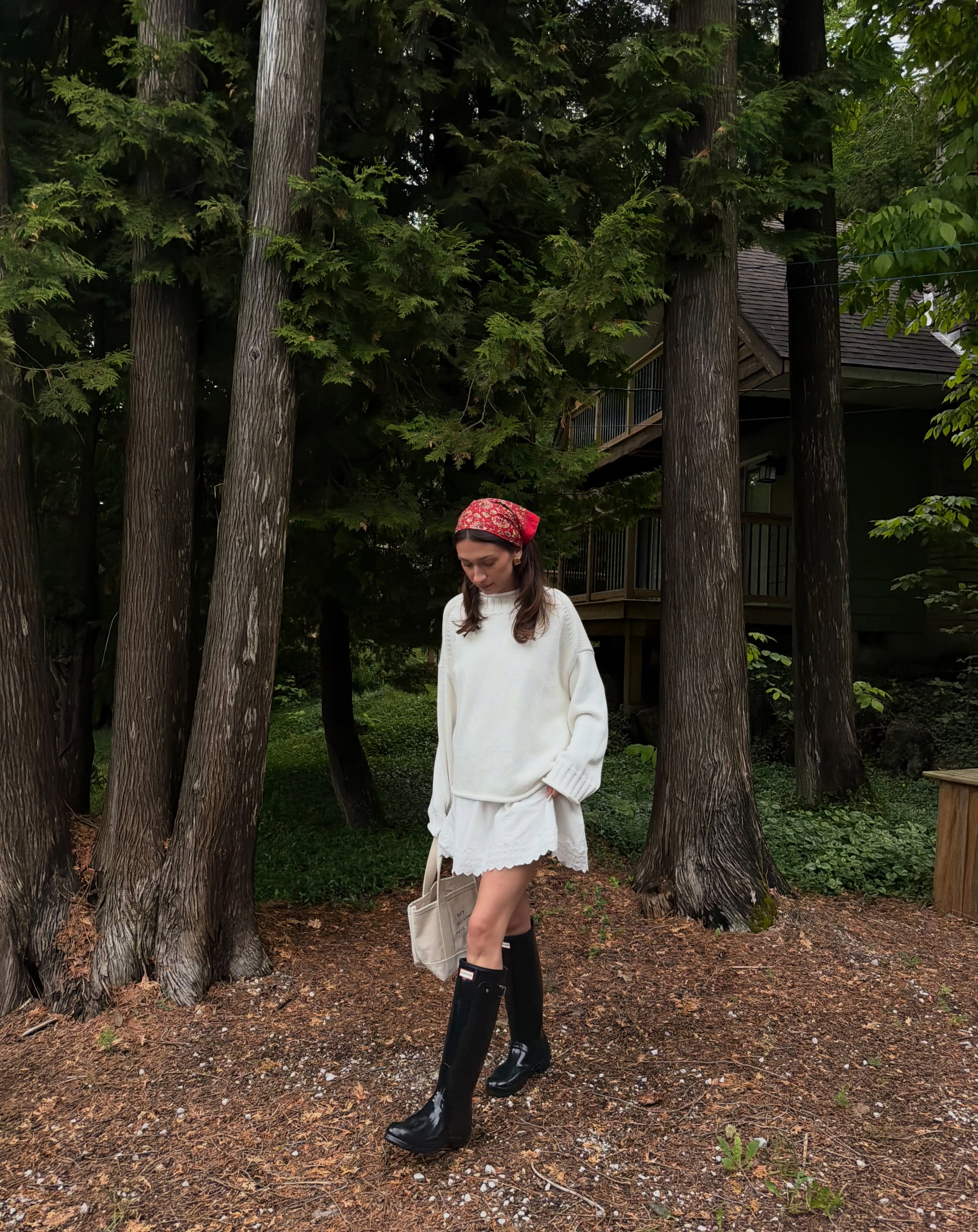 A young woman in a white outfit and black boots walks past tall trees in the foreground, with a rustic cabin visible in the wooded background.