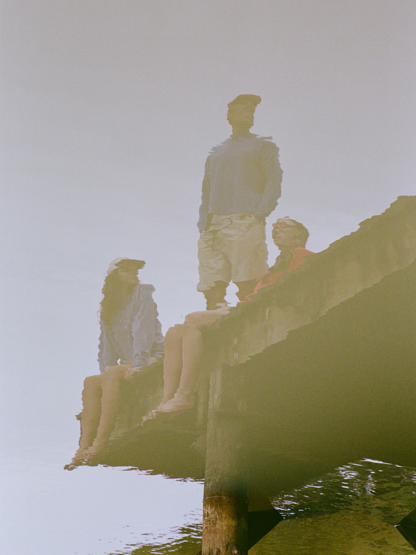 Three people sit and stand on a wooden pier against a hazy, pale sky.