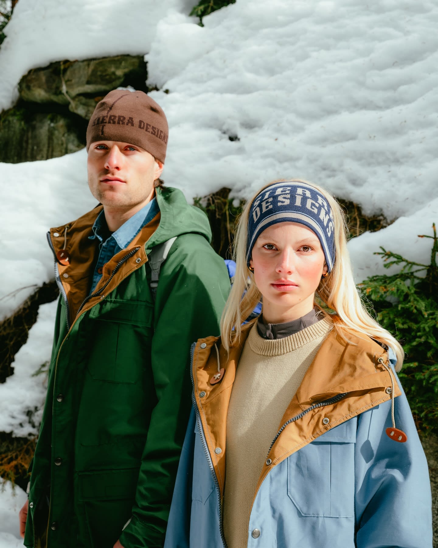 Two models in winter clothing stand in a snowy forest with rocks and trees in the background.