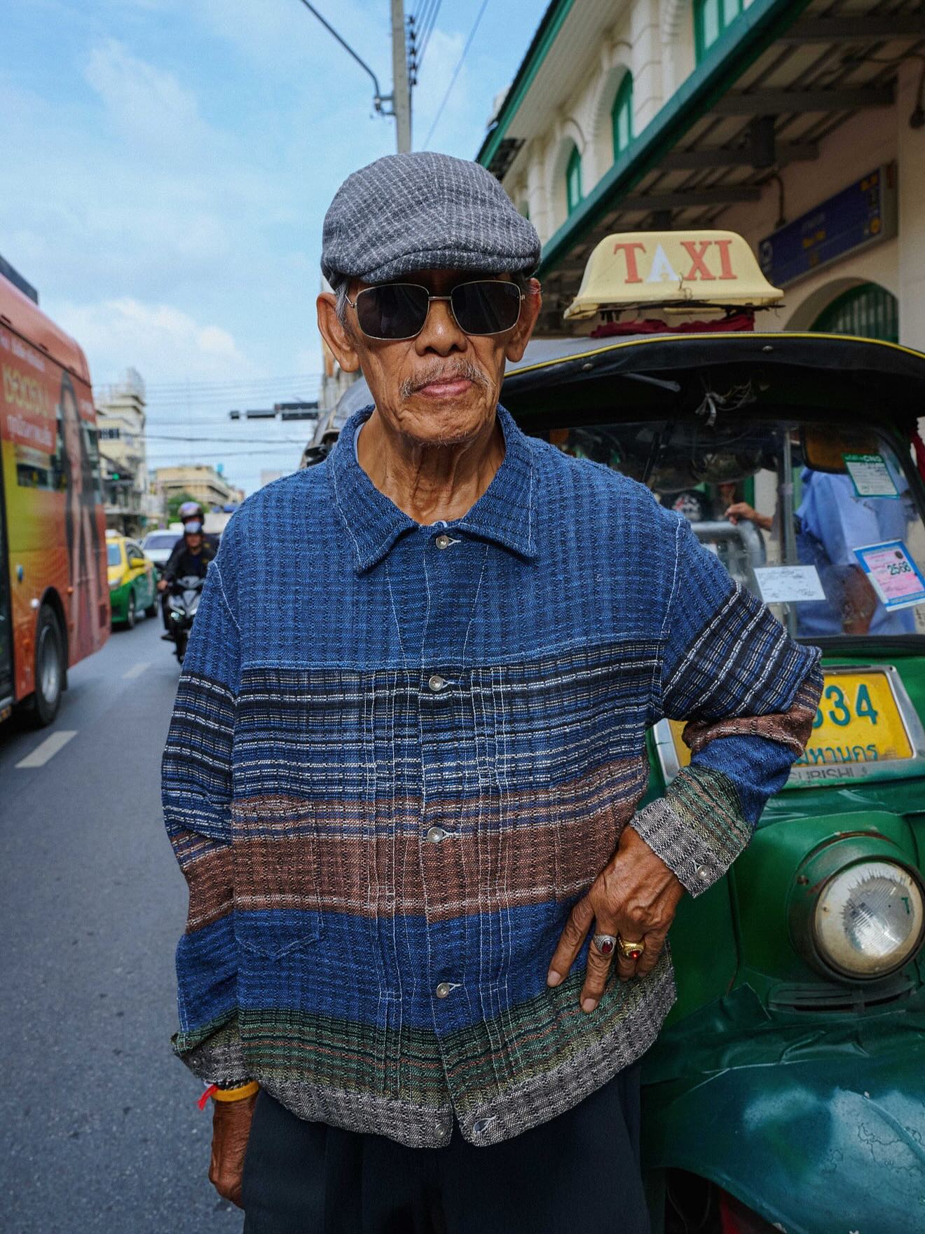 An elderly man in a flat cap and sunglasses stands in the foreground with a green tuk-tuk behind him, while a busy street with a bus and other vehicles forms the background.