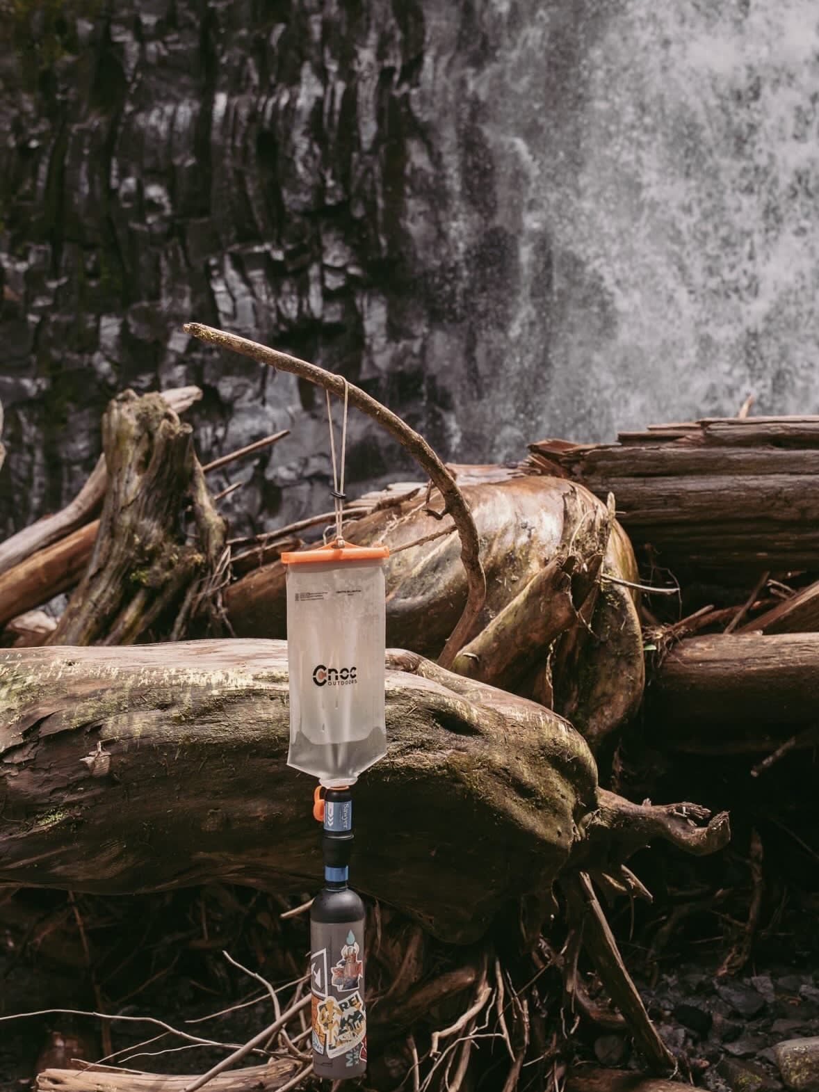 A water filtration bag hangs from a branch over fallen logs in front of a cascading waterfall.