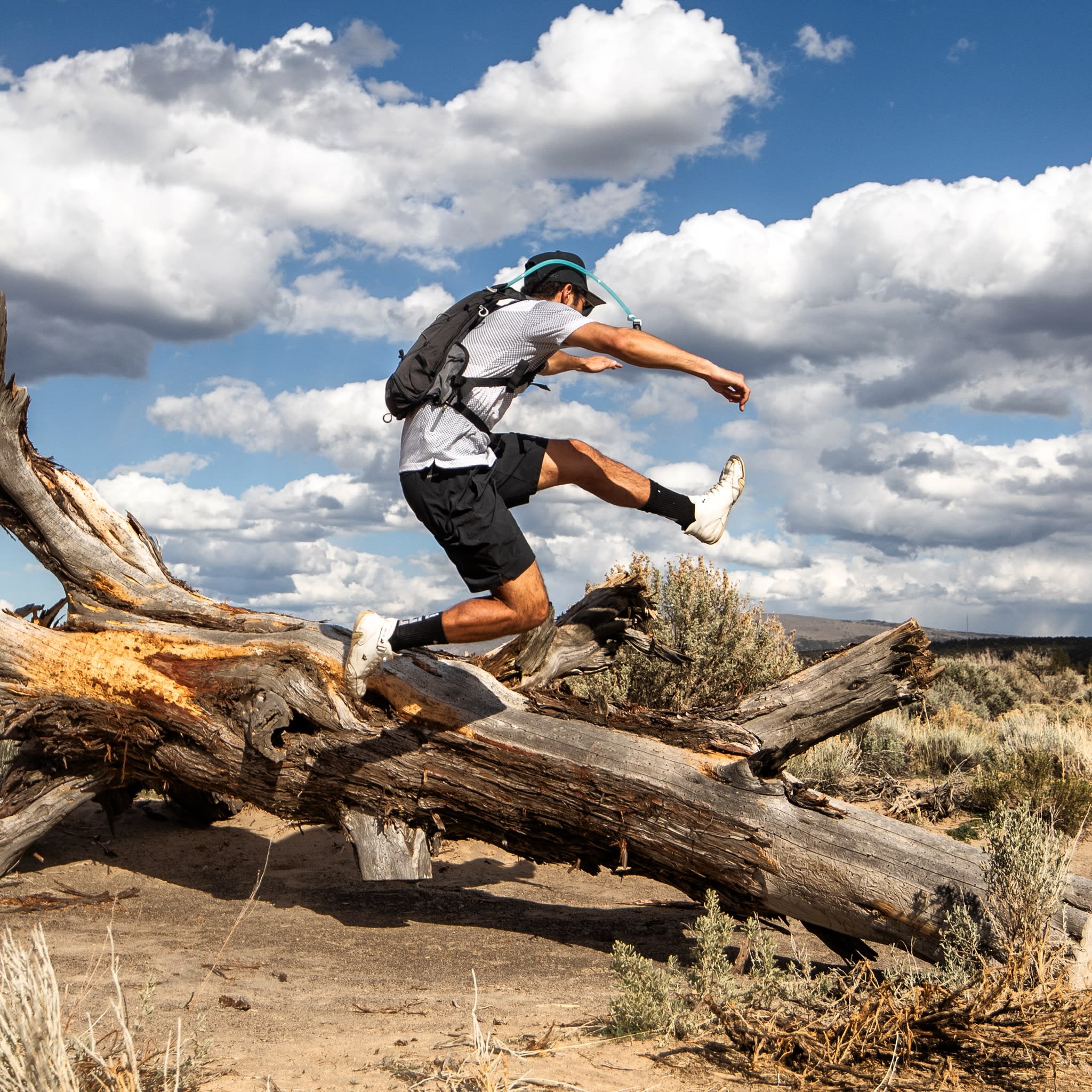 A man in athletic gear leaps across a large fallen log in a desert landscape under a bright, cloudy sky.