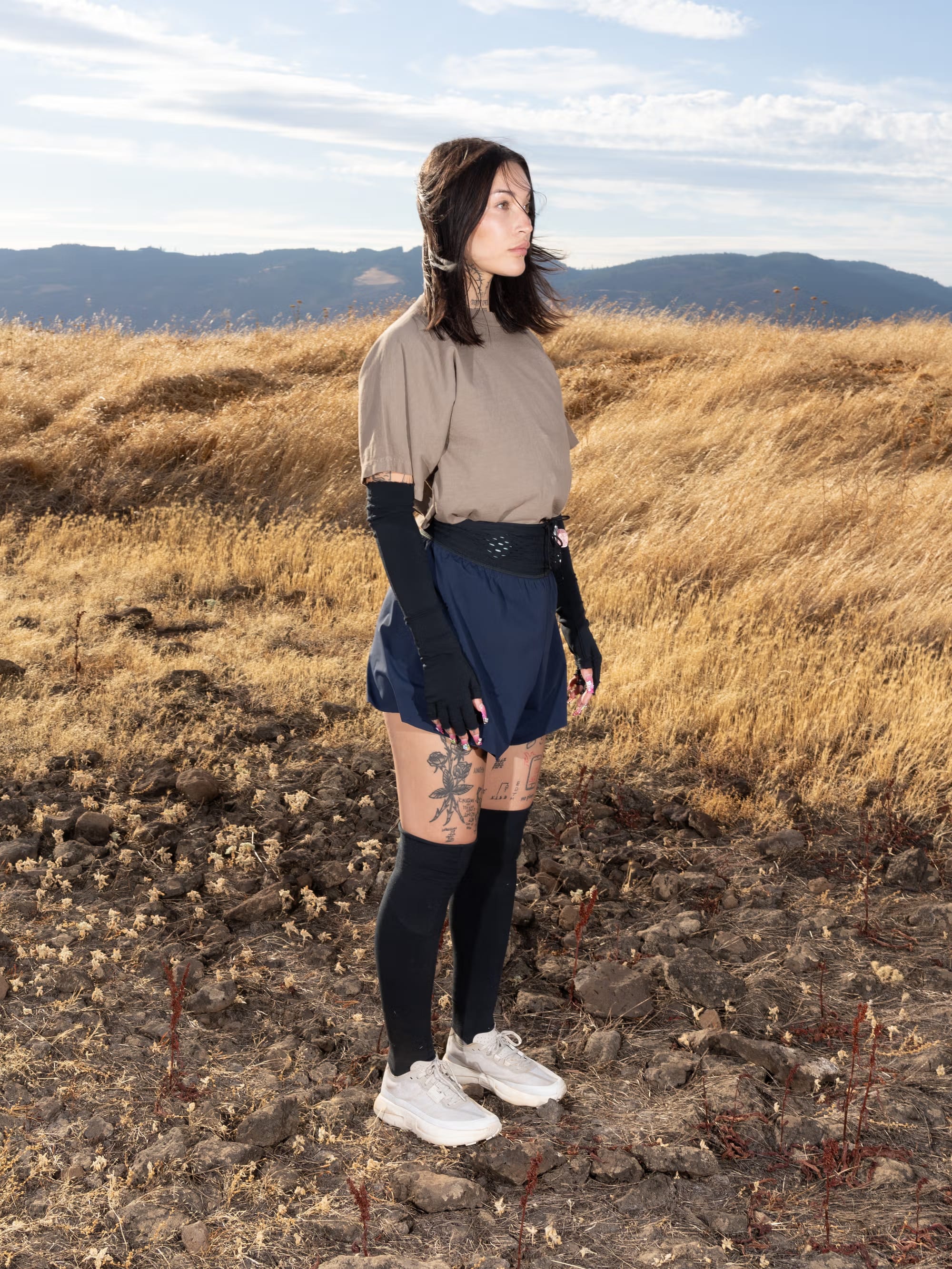 A woman wearing a tan shirt, blue shorts, and black accessories stands on rocky ground against a backdrop of golden fields and distant mountains.