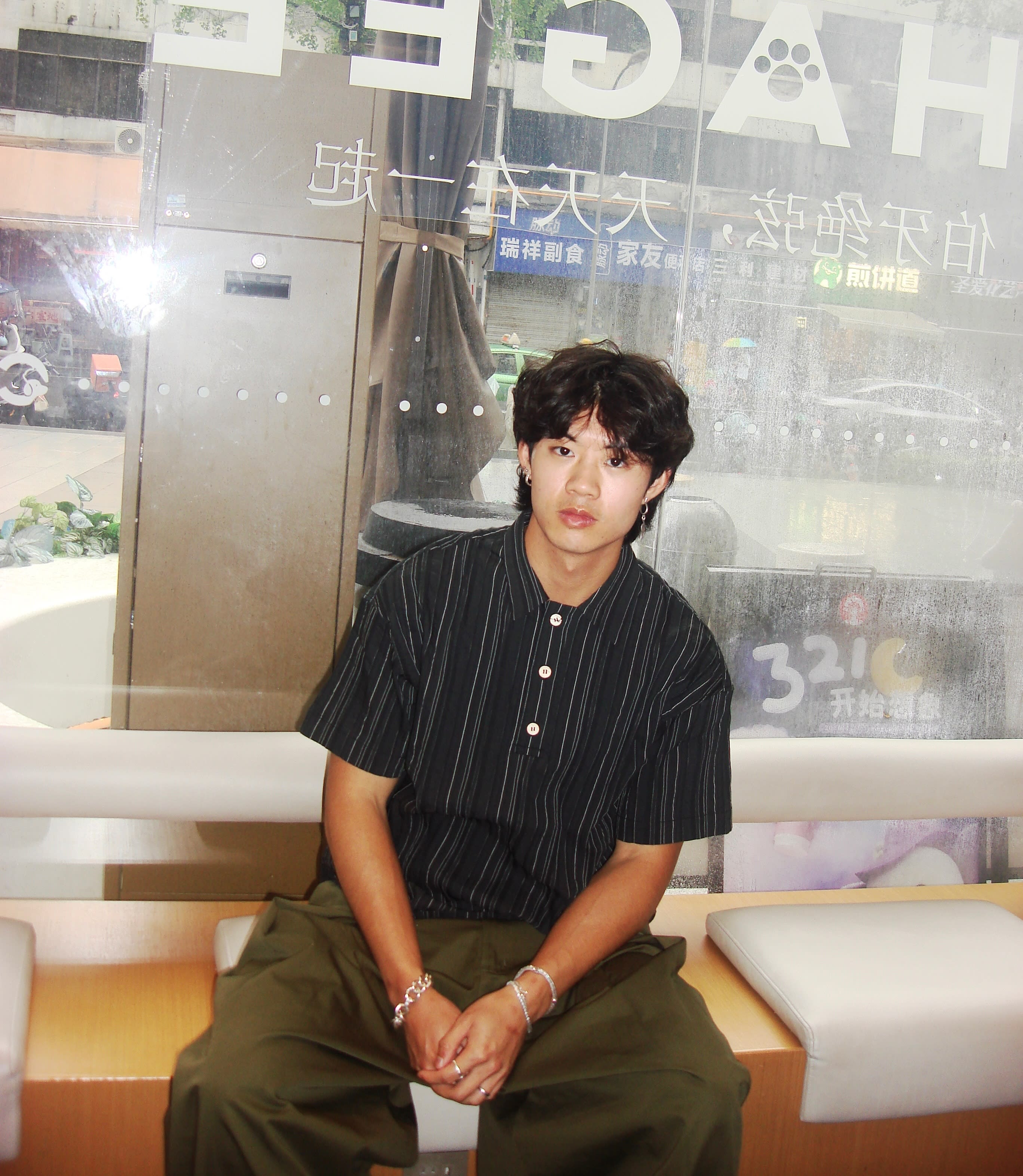 A young man in a striped black shirt and olive pants sits on a white bench in front of a glass window with Chinese text.