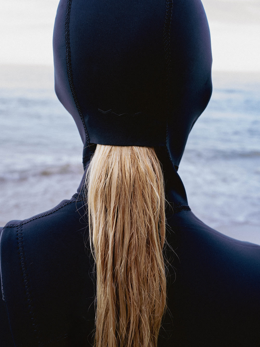 A person wearing a dark blue wetsuit hood with blonde hair flowing down the back stands before a blurred ocean backdrop.