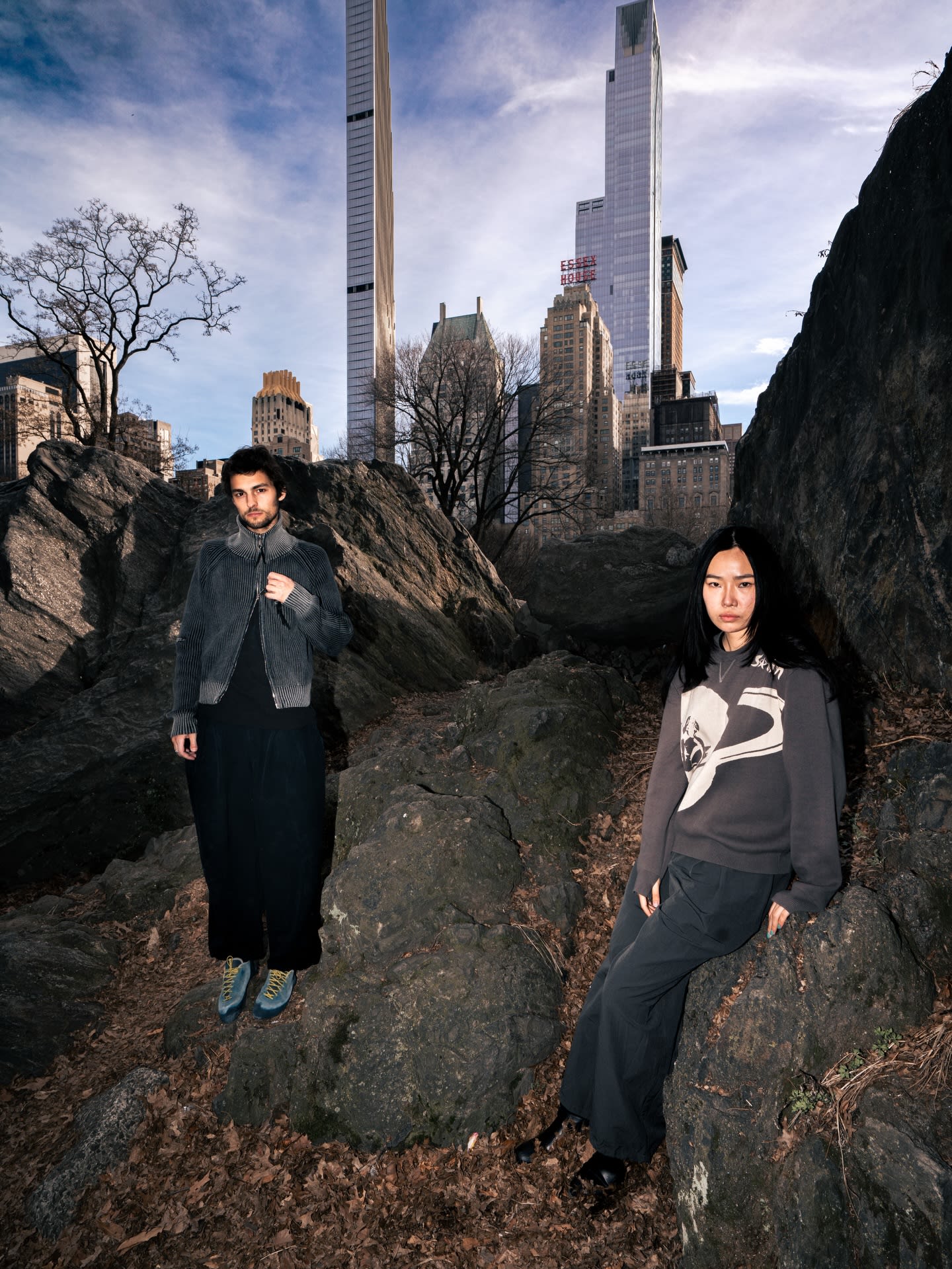 A man and a woman pose on rocky terrain in the foreground, with towering city skyscrapers rising into a blue sky in the background.