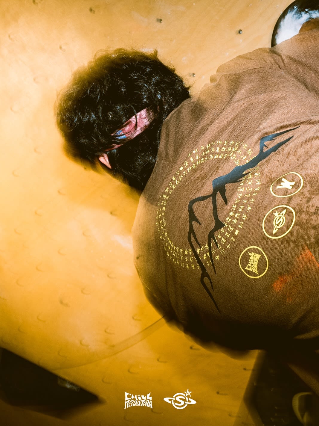 A person with dark curly hair wearing a brown graphic t-shirt leans against a textured yellow wall.