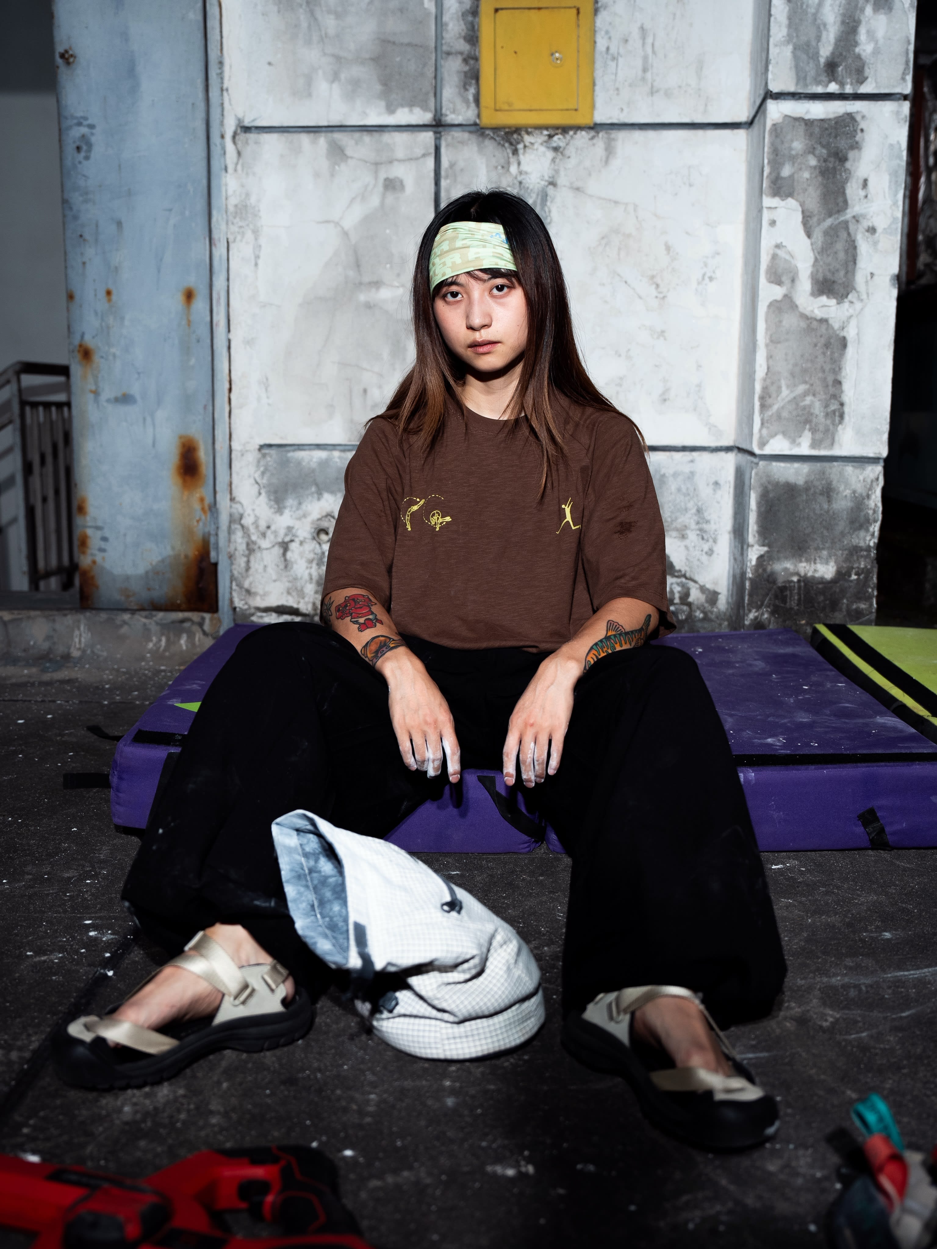 A woman with a green headband and tattoos sits on a purple crash pad in front of a weathered concrete wall.