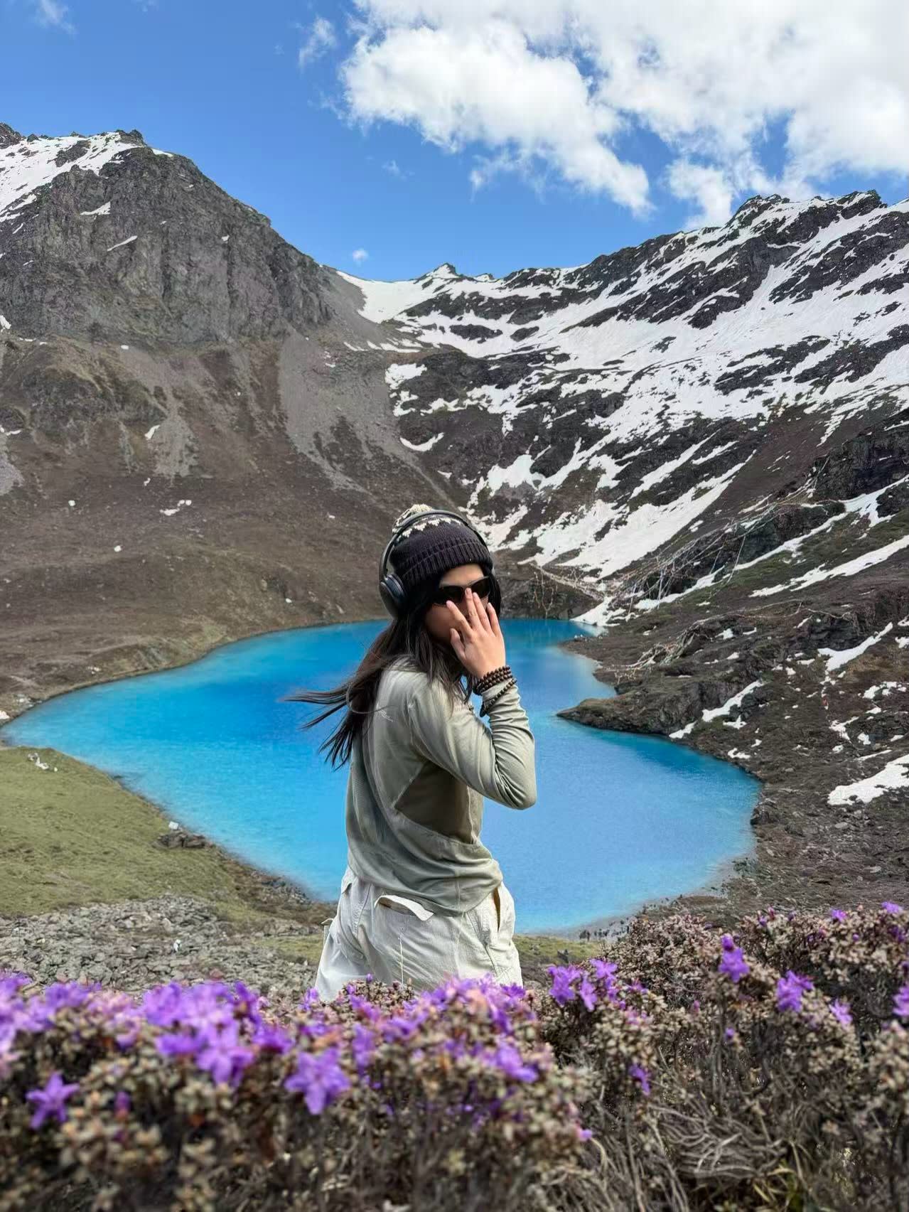 A woman wearing headphones and a beanie stands behind purple wildflowers in the foreground, with a bright blue alpine lake and snow-capped mountains in the background.