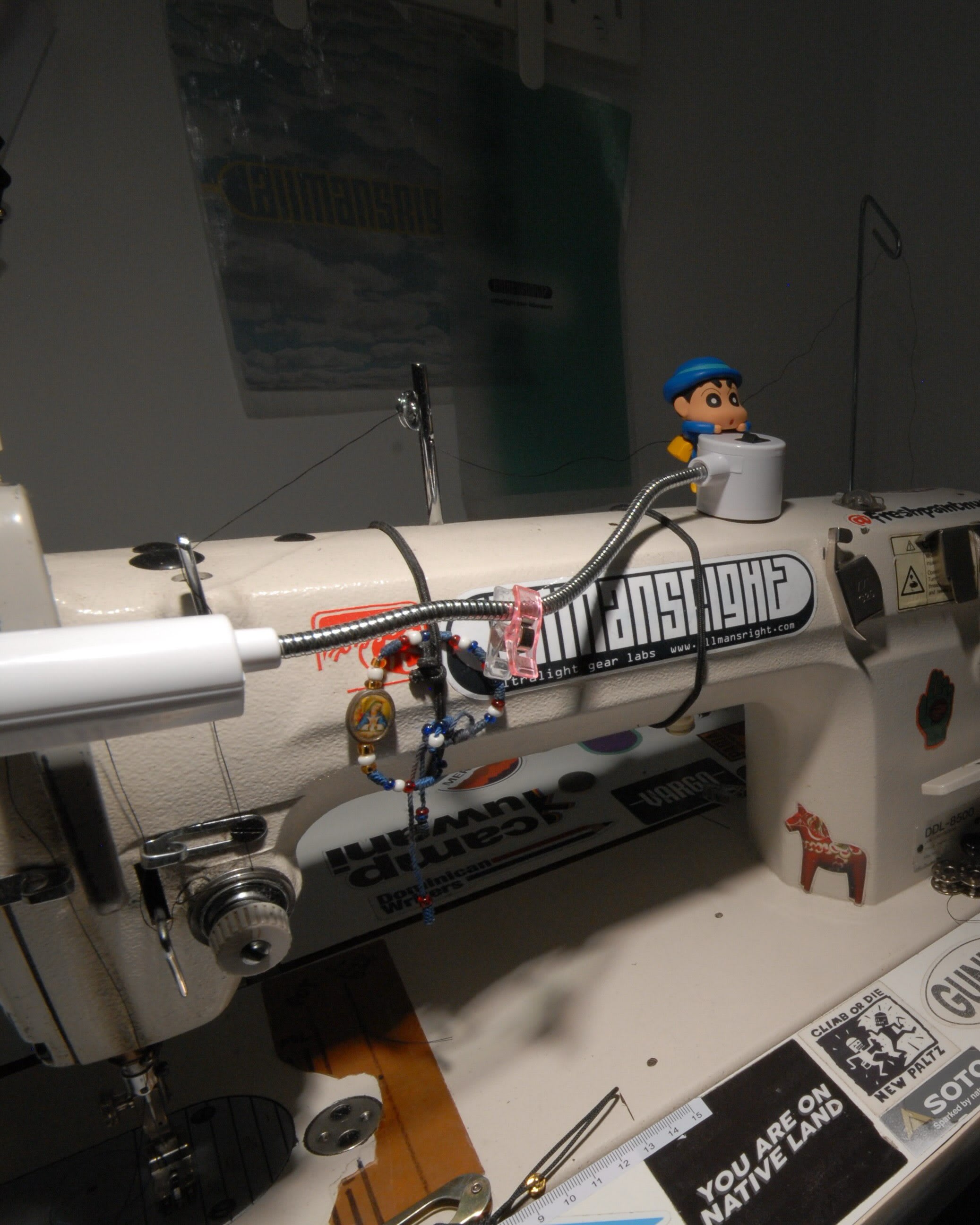 A small toy figure sits atop a sewing machine in the foreground, with a blurred background of what appears to be a workshop.