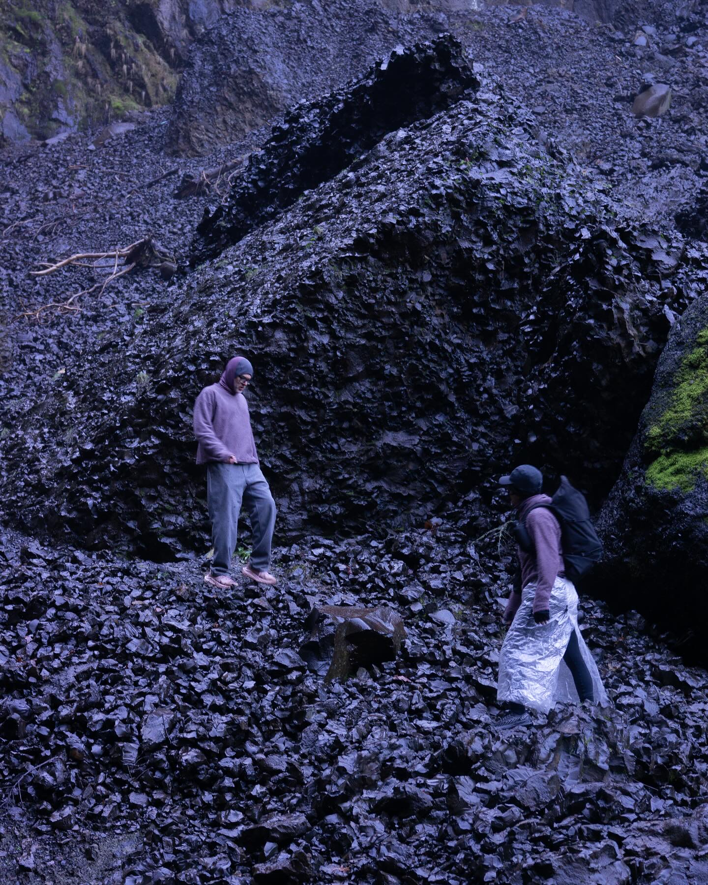 Two people stand on a slope of dark, jagged volcanic rocks against a backdrop of a massive, dark rock formation and a steep hillside.