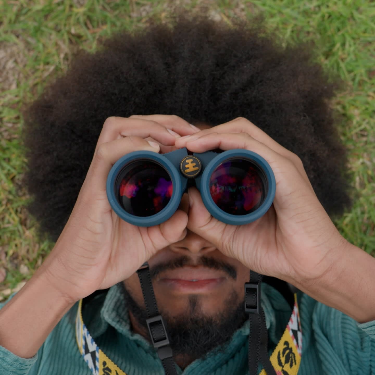 A person with an afro looks through binoculars in the foreground, with a grassy background.