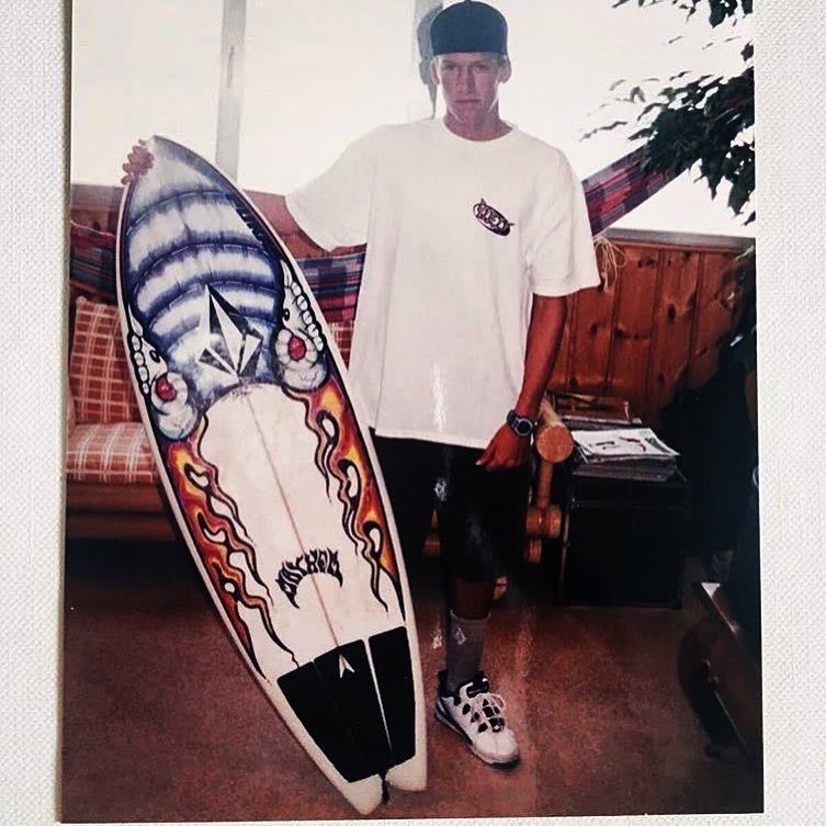 A man in a white t-shirt and black cap stands holding a colorful surfboard in a room with a couch and a plant.