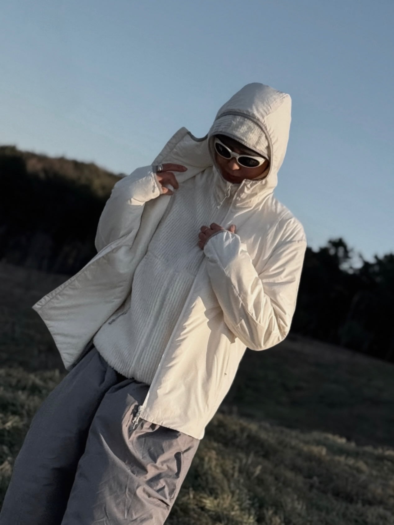 A person wearing a white hooded jacket, sunglasses, and grey pants stands in a grassy field against a backdrop of distant hills and a clear sky.