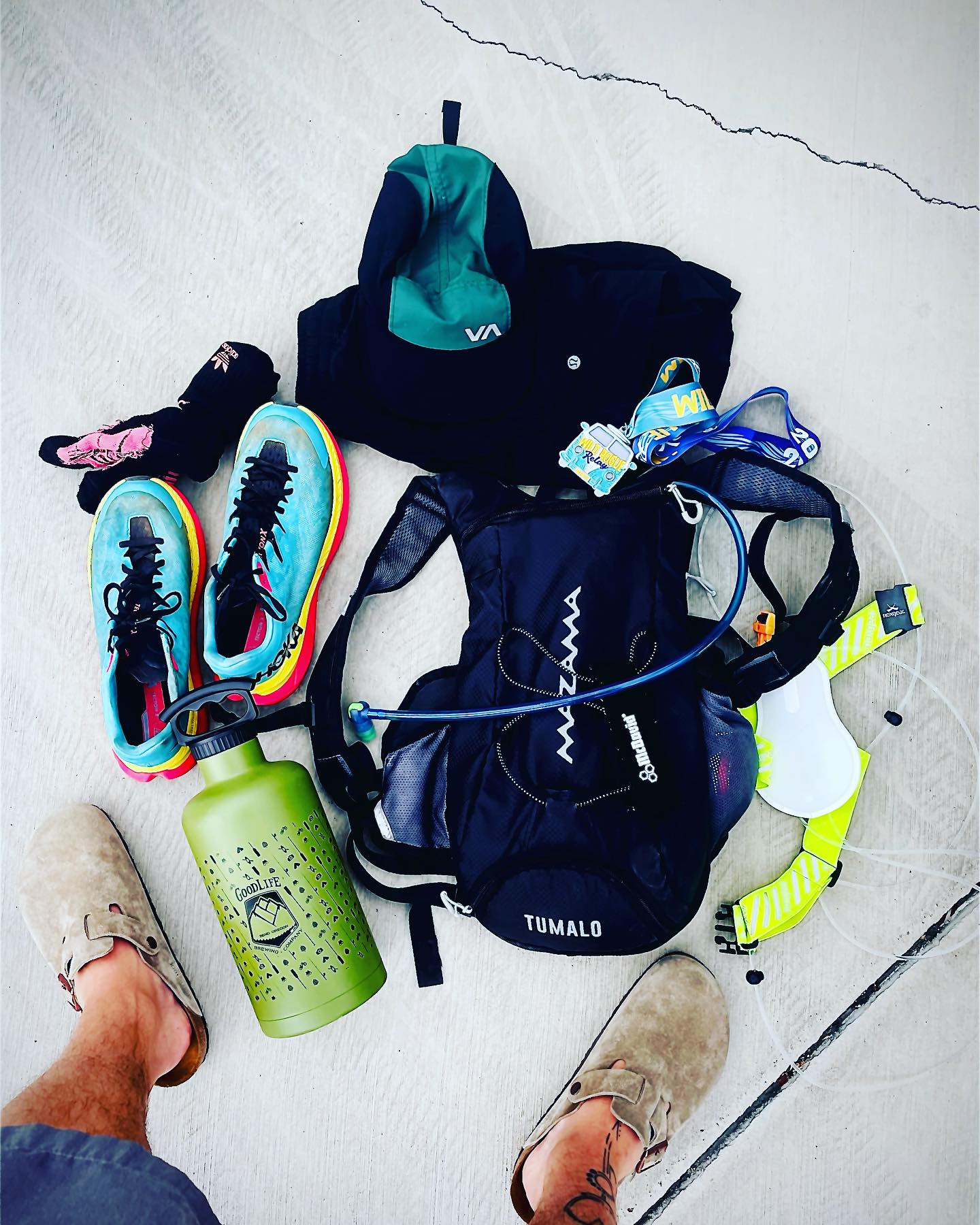 A pair of feet in tan slip-on shoes stands before a collection of hiking gear, including colorful sneakers, a hydration pack, a green water bottle, and a cap, all laid out on a light-colored concrete surface.