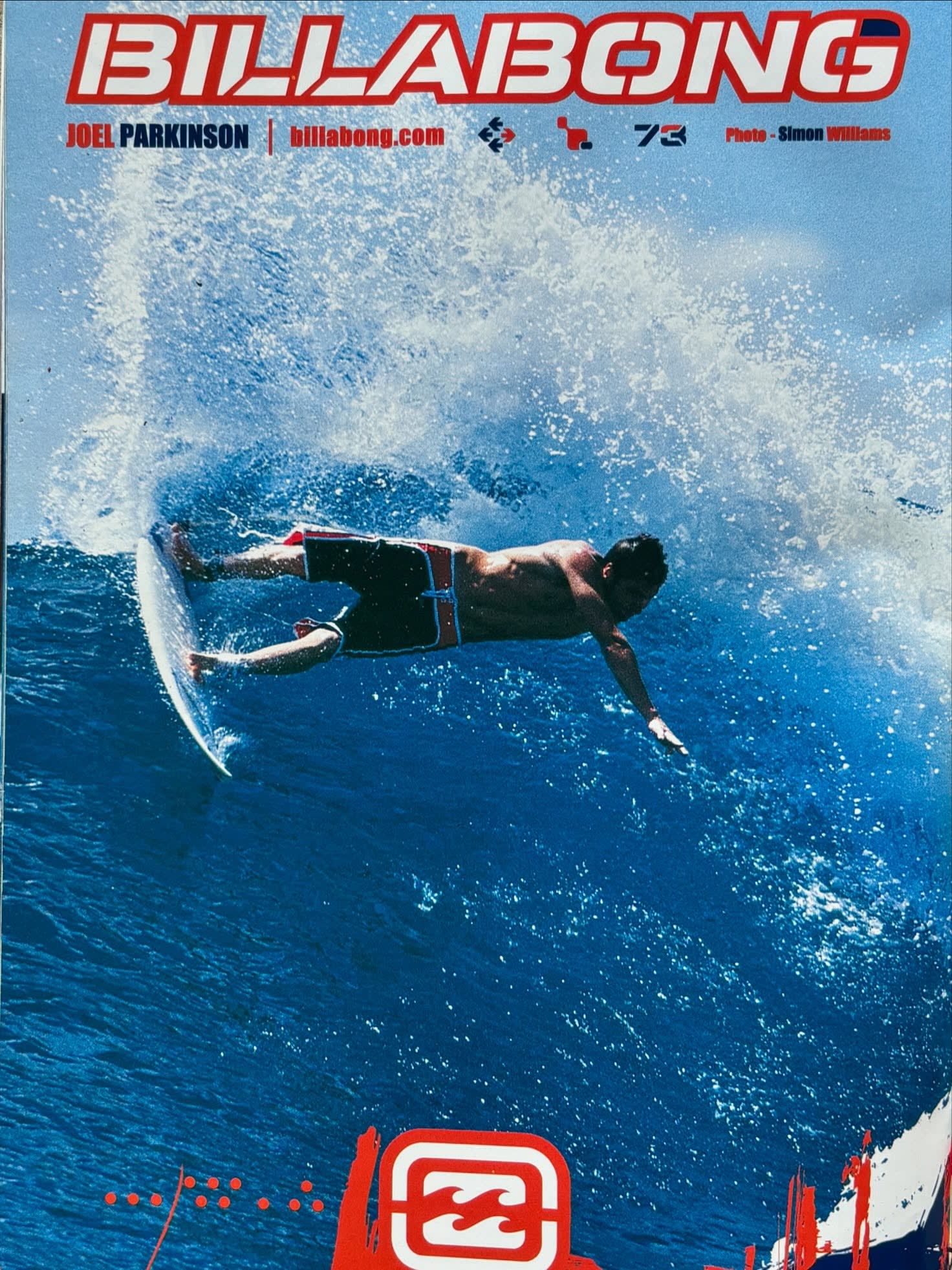 A surfer rides a white surfboard across a crashing blue wave under a clear sky.