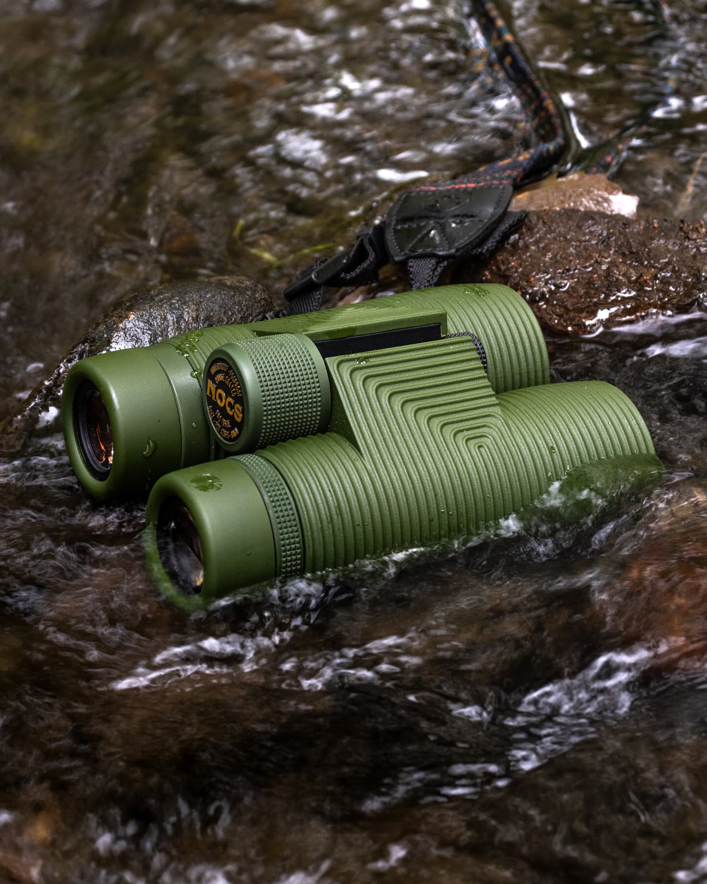 Green binoculars rest in the shallow, rippling water of a stream, with rocks and a strap visible in the background.